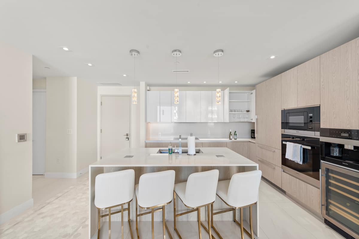 The open kitchen with a long island, four bar stools, a wine fridge and a cluster of pendant lights