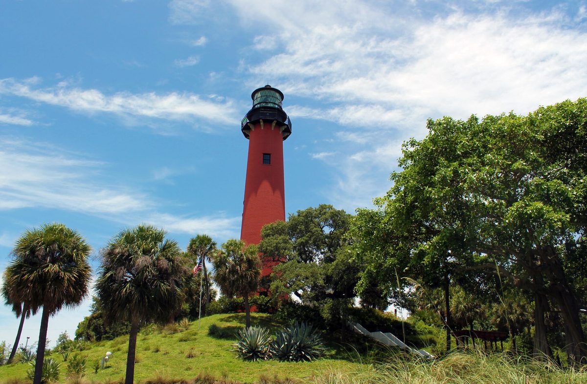 The red Jupiter Inlet Lighthouse on its grassy hill among palms and live oaks under a blue sky