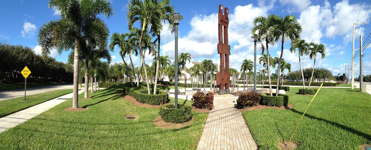 A palm-lined brick walkway and lawn leading to a tall red sculpture in Palm Beach Gardens