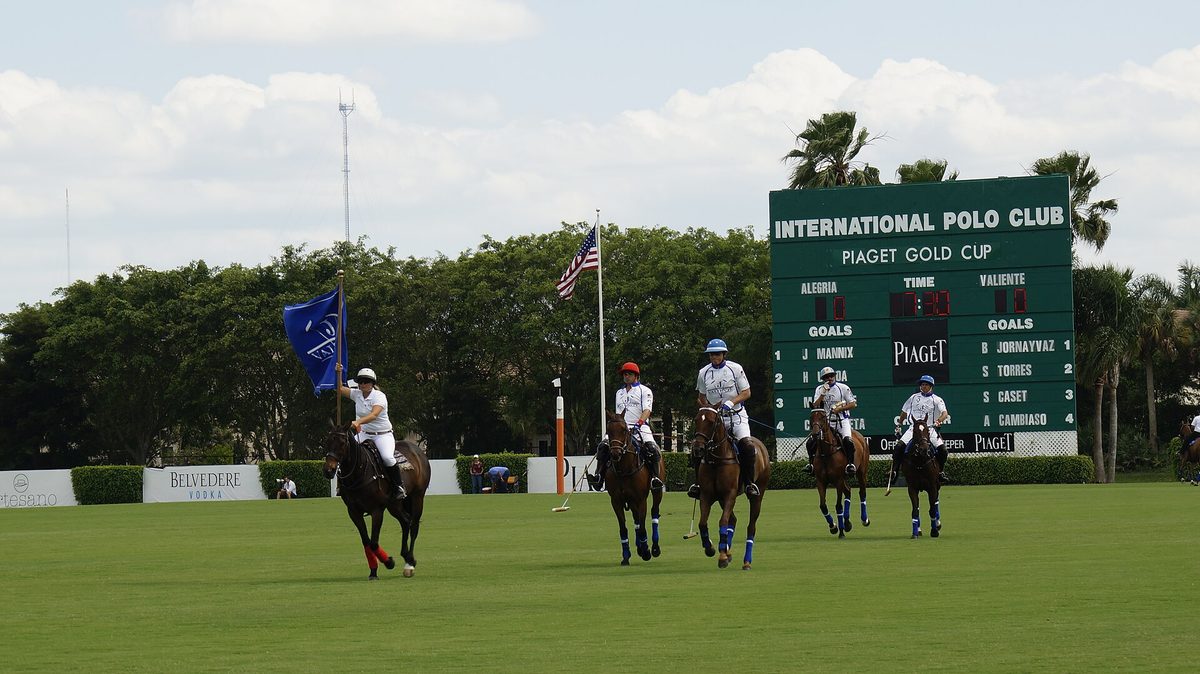 Polo players ride onto the field in Wellington, Florida, one carrying a team flag, in front of the Gold Cup scoreboard