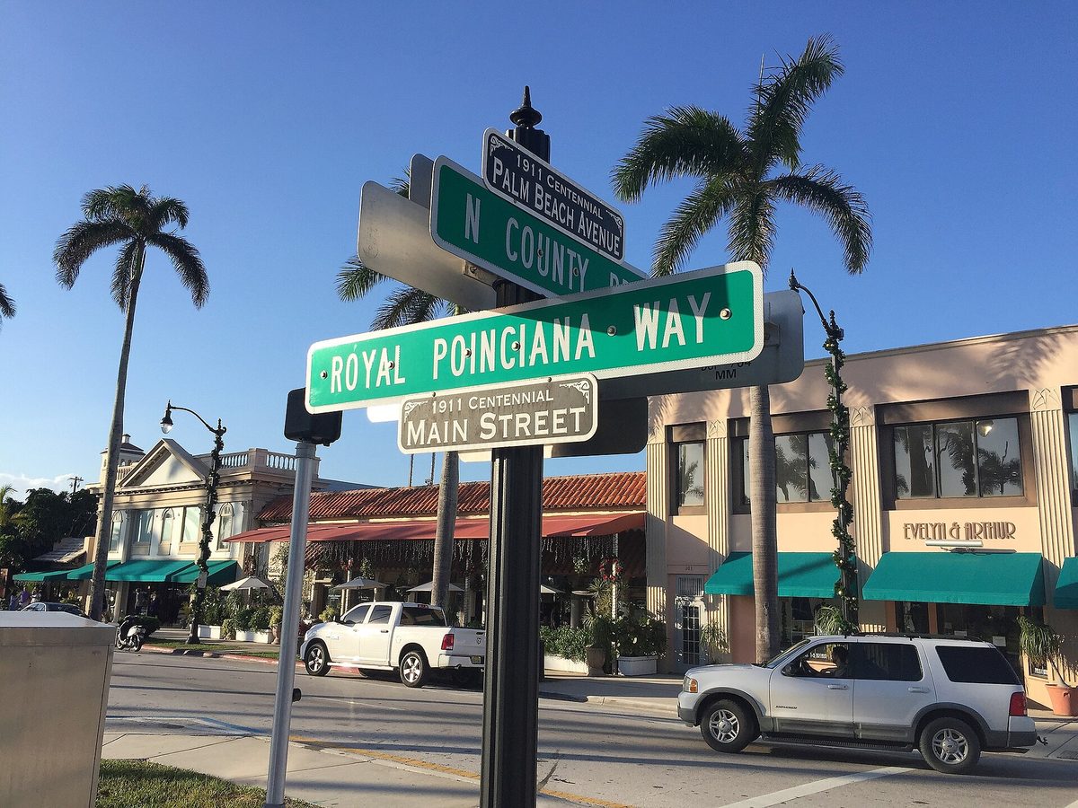 Street signs at Royal Poinciana Way and North County Road in Palm Beach, with palms and awninged shops behind