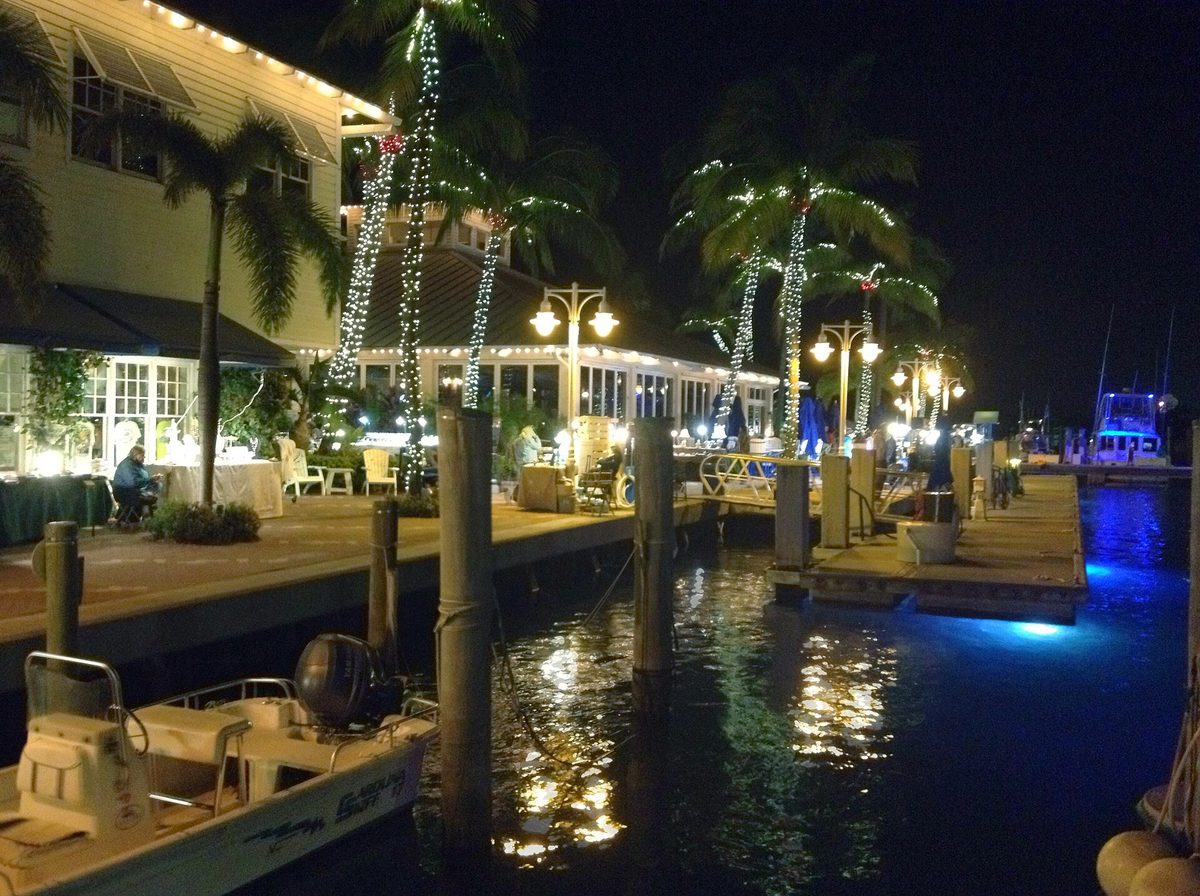 Sailfish Marina at night, with palm trees wrapped in white lights along the waterfront dining terrace and boats at the dock