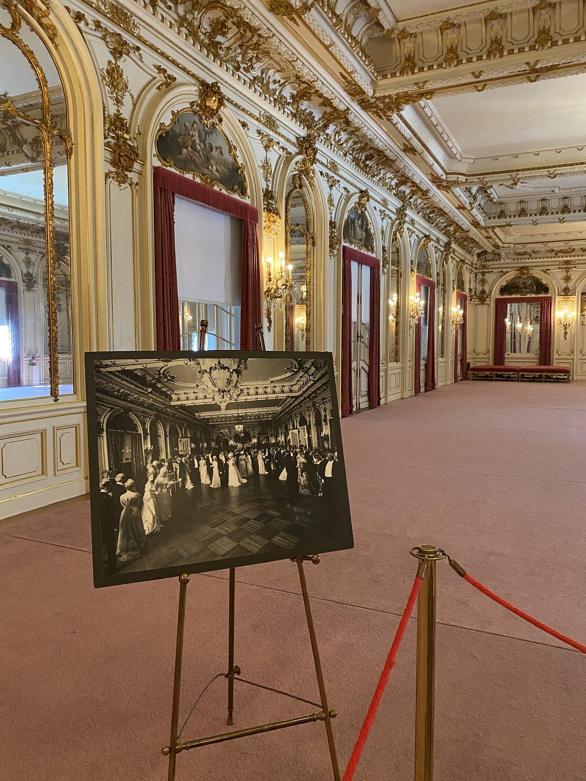 The gilded ballroom at Whitehall with mirrors, red drapes and a historic photograph of a ball on an easel