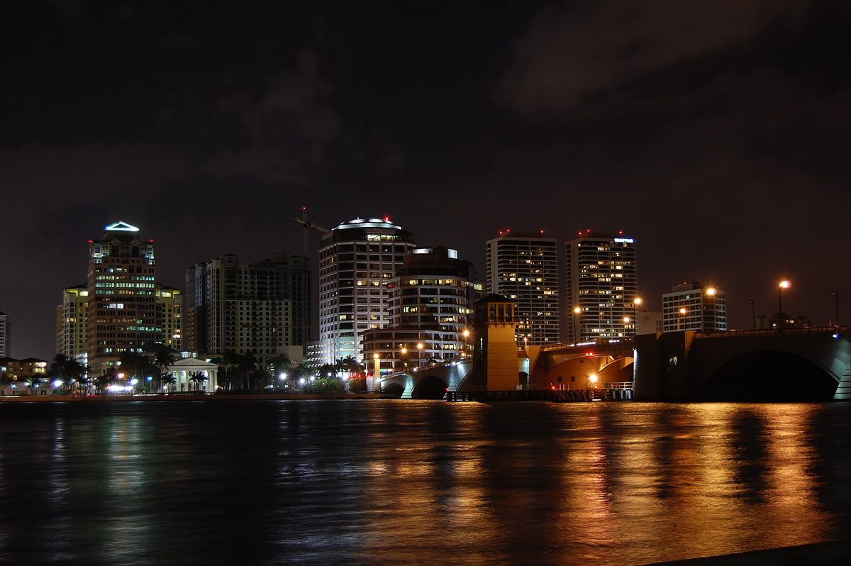 The downtown West Palm Beach skyline at night across the Intracoastal, its lights reflected in the water beside a bridge
