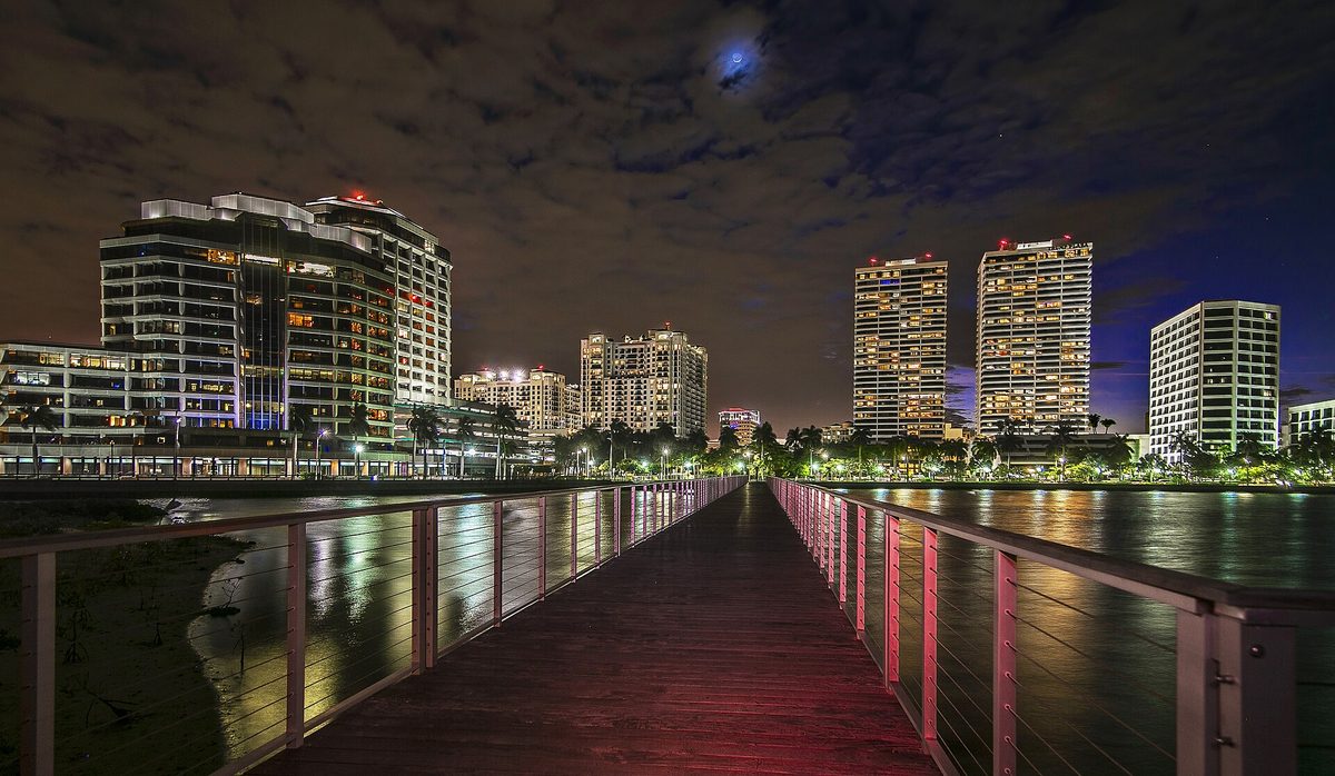 A boardwalk pier over the water at night, with the lit towers of downtown West Palm Beach along the waterfront